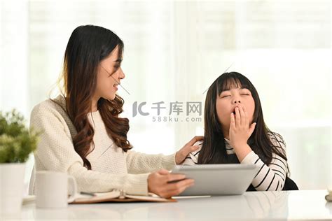 昏昏欲睡的小女孩在家里和妈妈一起做作业时打哈欠、用手捂嘴的照片高清摄影大图-千库网