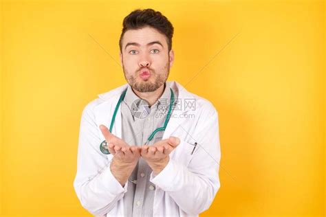 年轻的高加索女医生在黄色背景下用嘴唇吹口吻握着手掌来发出空气之吻高清图片下载-正版图片504552010-摄图网