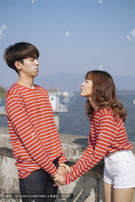 一对身穿红色t恤的年轻夫妇站在大坝上，手挽着手，卷曲的长发女孩试图亲吻她的男朋友。,摄影素材,汇图网www.huitu.com