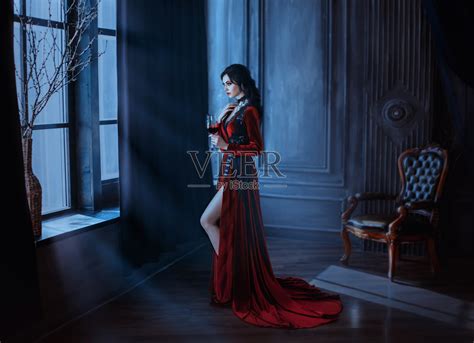 美丽的年轻性感的女人吸血鬼在中世纪黑暗的城堡。红色哥特式长裙。黑色的卷发。背景复古室内。把酒捧在酒杯里。形象女王的夜晚恐怖节日万圣节照片摄影 ...
