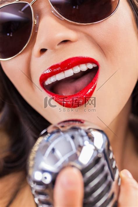 在夜总会里，戴着飞行员太阳镜、嘴巴红唇对着老式麦克风唱歌的漂亮女歌手的特写高清摄影大图-千库网